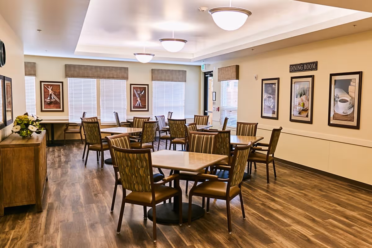 Bright senior living facility dining room with multiple tables and chairs, framed artwork, and wood flooring.
