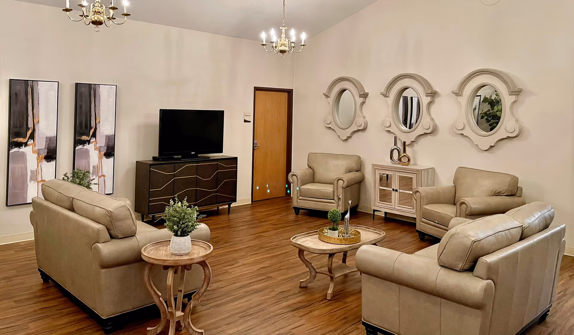 A cozy living room with two beige leather sofas, two matching armchairs, a wooden coffee table with decorative items, and a small round side table with a potted plant. The room features wooden flooring, three decorative mirrors on the wall, two abstract paintings, a flat-screen TV on a dark wooden cabinet, and two chandeliers hanging from the ceiling.