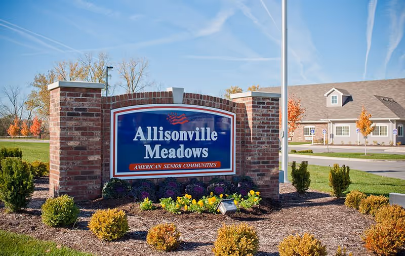 Brick monument entrance sign reading "Allisonville Meadows" surrounded by landscaping with the community building visible in the background.
