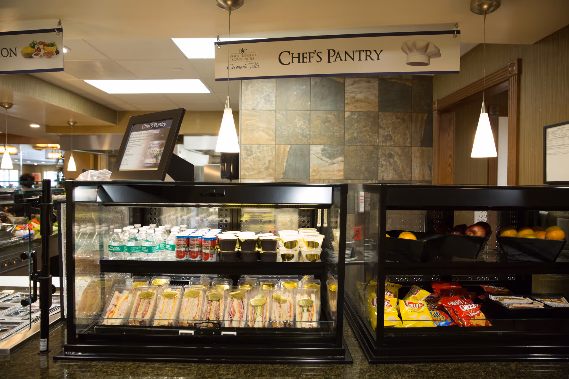 A countertop display case labeled "Chef's Pantry" holding bottled water, prepackaged sandwiches, snacks and fruit in a dining area.