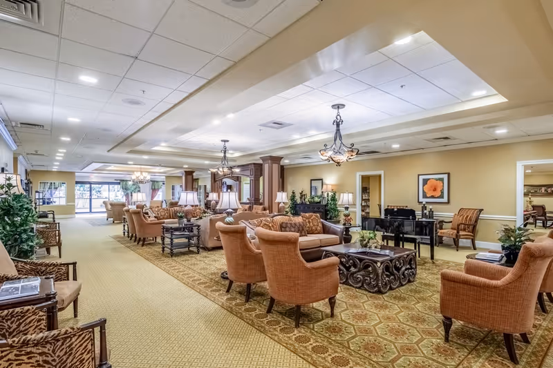 Spacious senior living facility common area with multiple seating arrangements including armchairs and sofas, decorative lamps, chandeliers, plants, and framed artwork on the walls. The room has a patterned carpet and a grand piano in the background.