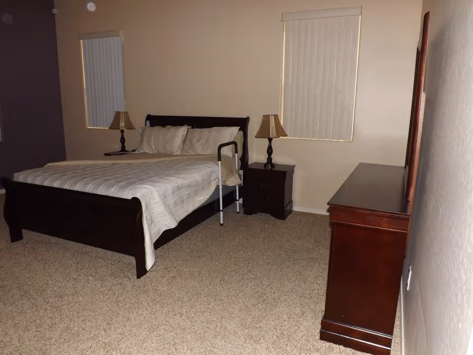 A bedroom with a dark wooden bed frame, beige bedding, two matching nightstands with lamps, beige carpet, and two windows with vertical blinds. There is a dark wooden dresser on the right side of the room.