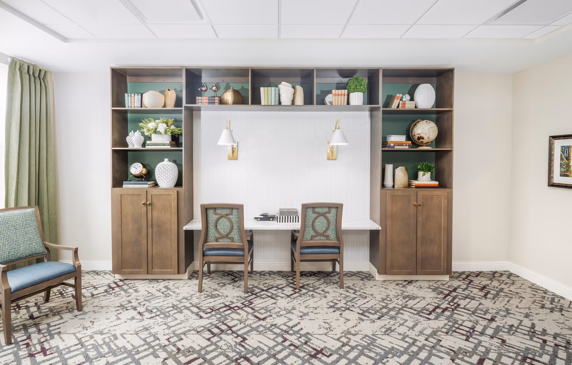 A well-lit interior room featuring a built-in wooden shelving unit with decorative items such as books, vases, a globe, and plants. Below the shelves is a white desk with two chairs, each with a patterned backrest. The room has a patterned carpet, light-colored walls, a framed picture on the right wall, and a green curtain on the left side.