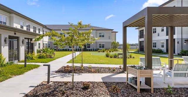 Outdoor courtyard area at Highpoint at Stonecrest featuring a paved walkway, young trees, landscaped garden beds with mulch, and a shaded seating area with chairs and a small table. The courtyard is surrounded by two-story residential buildings under a clear blue sky.