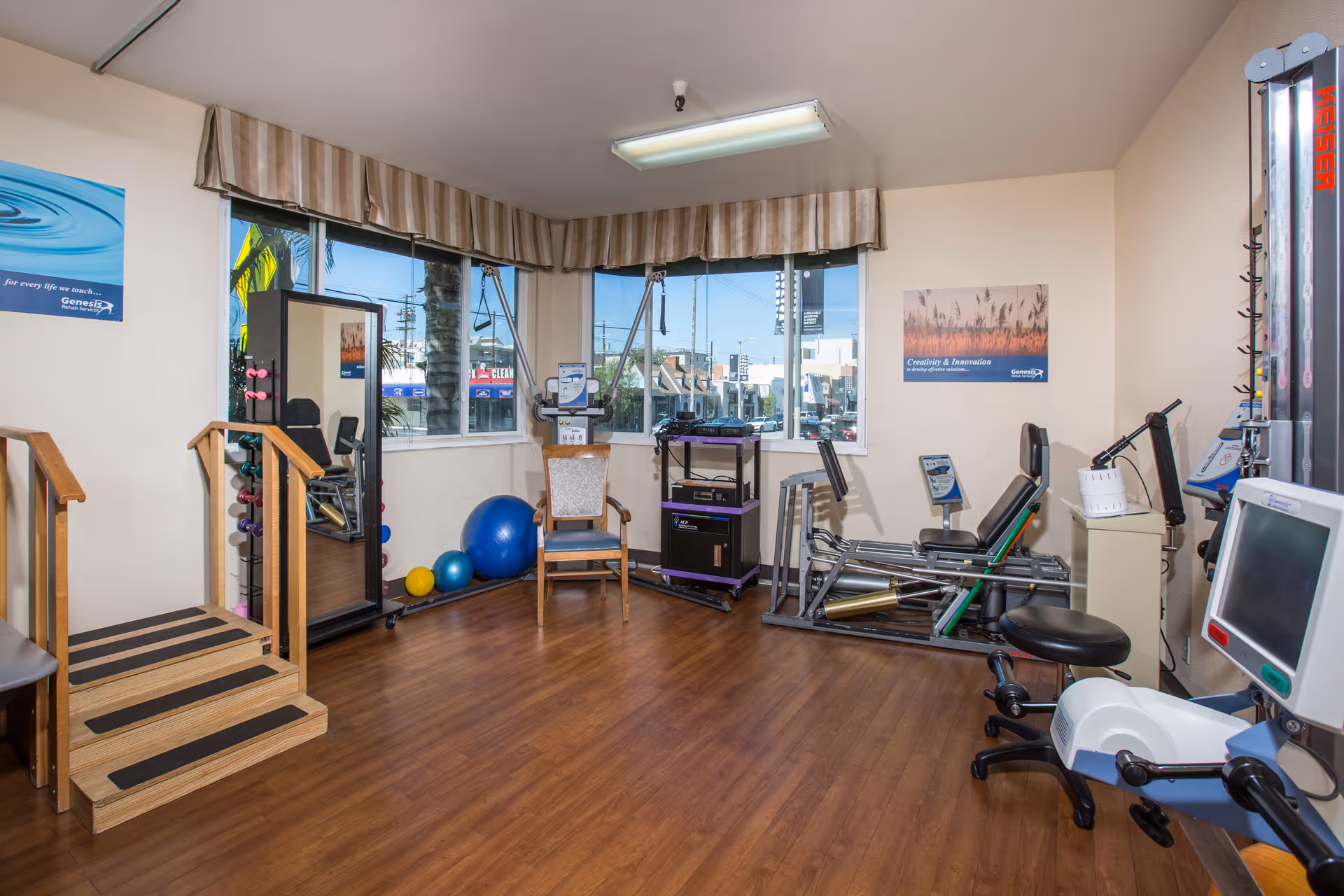 A rehabilitation or physical therapy room with exercise equipment including a leg press machine, an arm bike, therapy balls, a mirror, and a small set of stairs with handrails. The room has large windows with striped valances, wood flooring, and motivational posters on the walls.