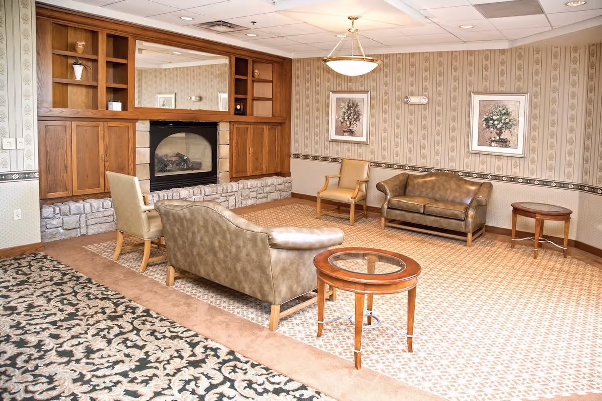 A senior living facility lounge area with patterned carpet and wallpaper. The room features a stone and wood fireplace with built-in shelves and cabinets, a large mirror above the fireplace, two leather sofas, two armchairs, two round wooden side tables with glass tops, and framed floral artwork on the walls. A ceiling light fixture hangs in the center of the room.