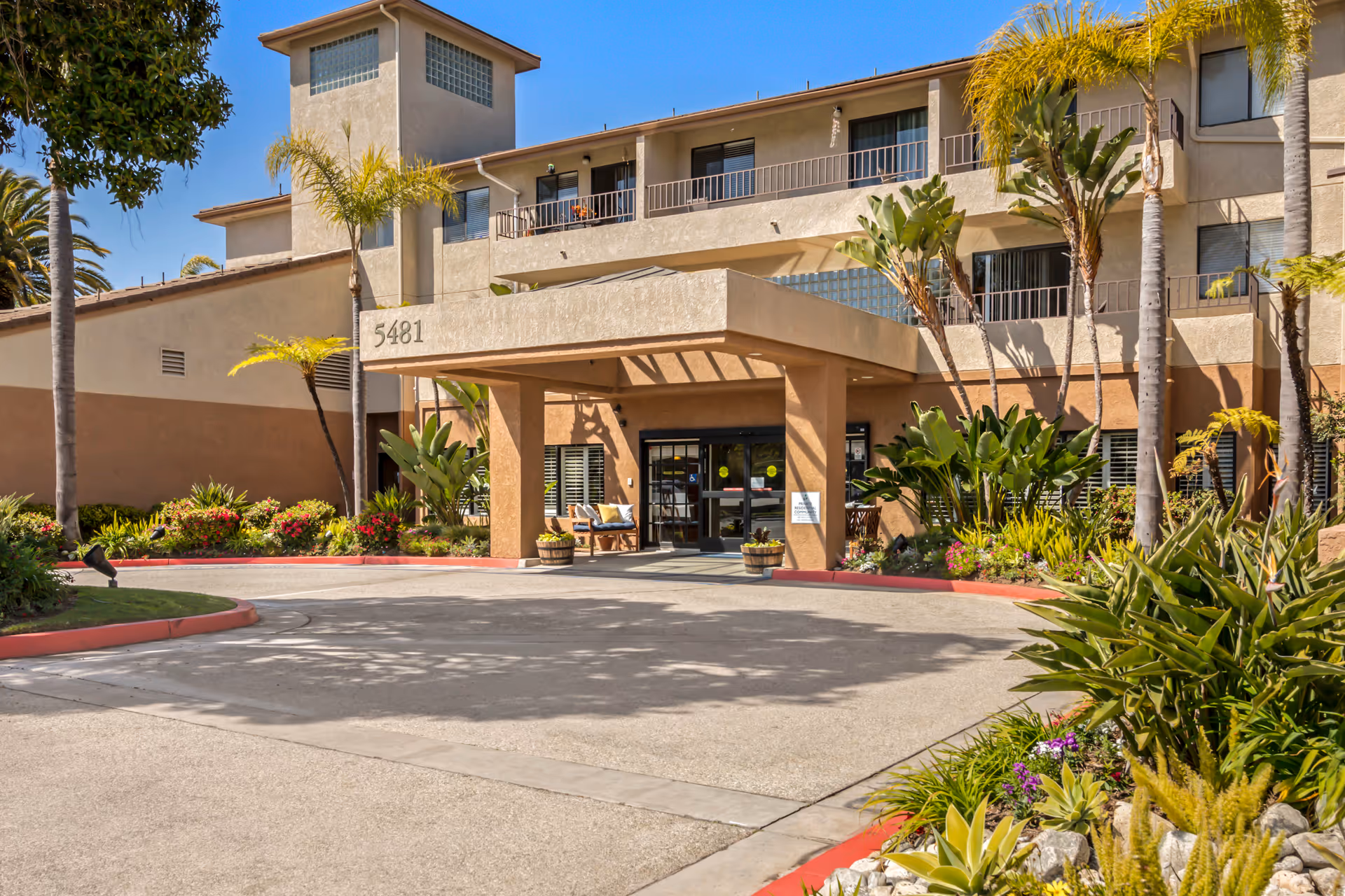 Exterior view of the Brookdale South Bay senior living facility showing the main entrance with a covered drop-off area, surrounded by palm trees and landscaped greenery under a clear blue sky.