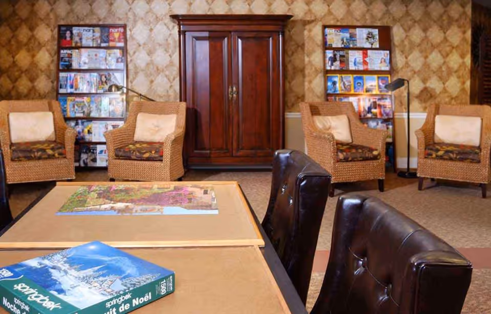 A cozy communal lounge with wicker armchairs, leather chairs, magazine racks, a puzzle table, and a wooden cabinet against patterned wallpaper.