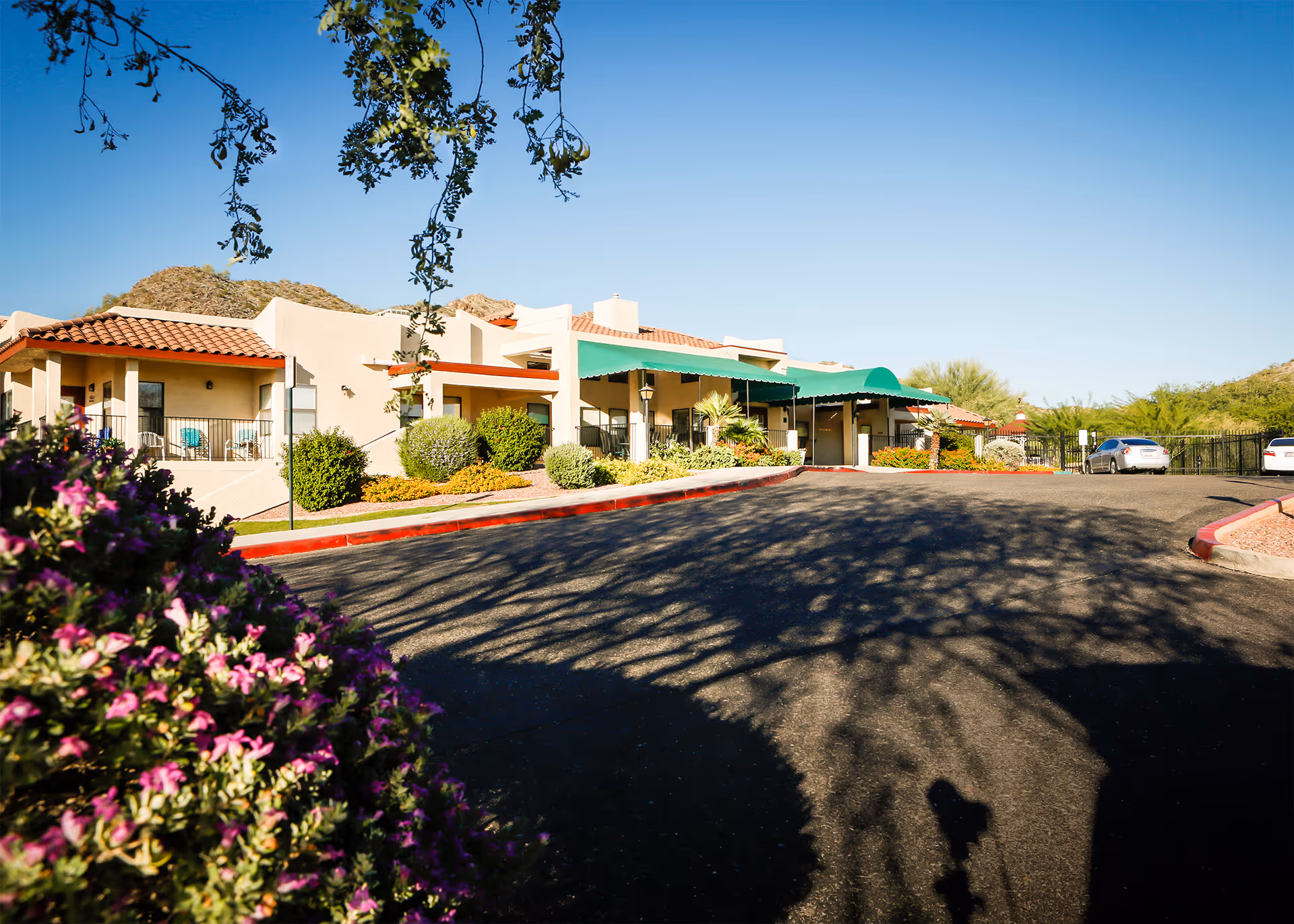 Exterior view of a senior living facility with beige stucco buildings, red tile roofs, green awnings, and a paved driveway. There are bushes and flowering plants in the foreground and mountains in the background under a clear blue sky.