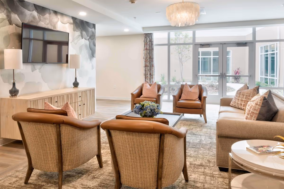 A bright and modern living room in an assisted living facility featuring a beige sofa with patterned pillows, four armchairs with leather and fabric upholstery arranged around a coffee table with a floral centerpiece. A flat-screen TV is mounted on a wall with a gray and white abstract design, flanked by two table lamps on a wooden console. Large windows and glass doors provide natural light and a view of an outdoor courtyard.