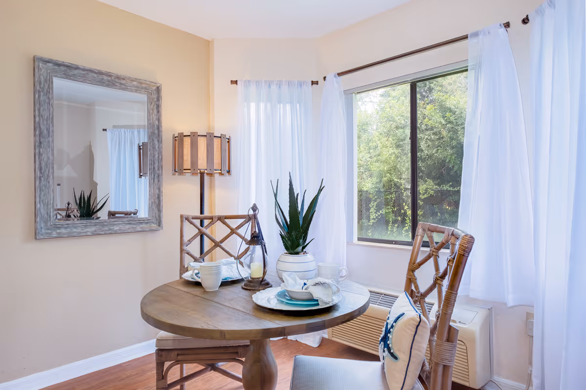 A cozy dining area with a round wooden table set for two, featuring plates, cups, and a small plant centerpiece. Two wooden chairs with cushions are placed around the table. A large window with white curtains allows natural light to brighten the room, and a decorative mirror and floor lamp are visible on the beige walls.