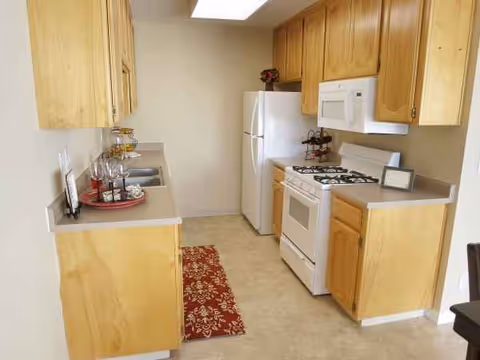 Narrow kitchen with wooden cabinets, white appliances, and a red rug in a senior living facility.