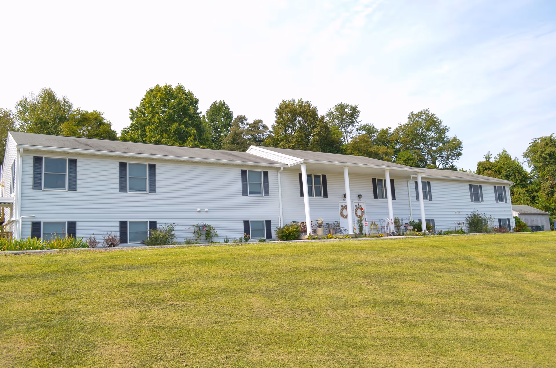 Country Acres Assisted Living's single-story white building with a front entrance and columns sits on an expansive grassy lawn.