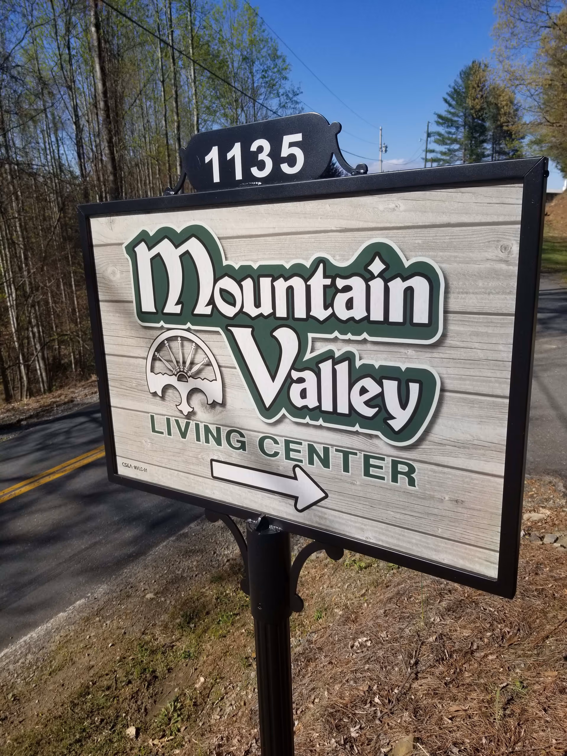Outdoor sign for Mountain Valley Living Center with the address number 1135 displayed above it, situated next to a road with trees and clear blue sky in the background.