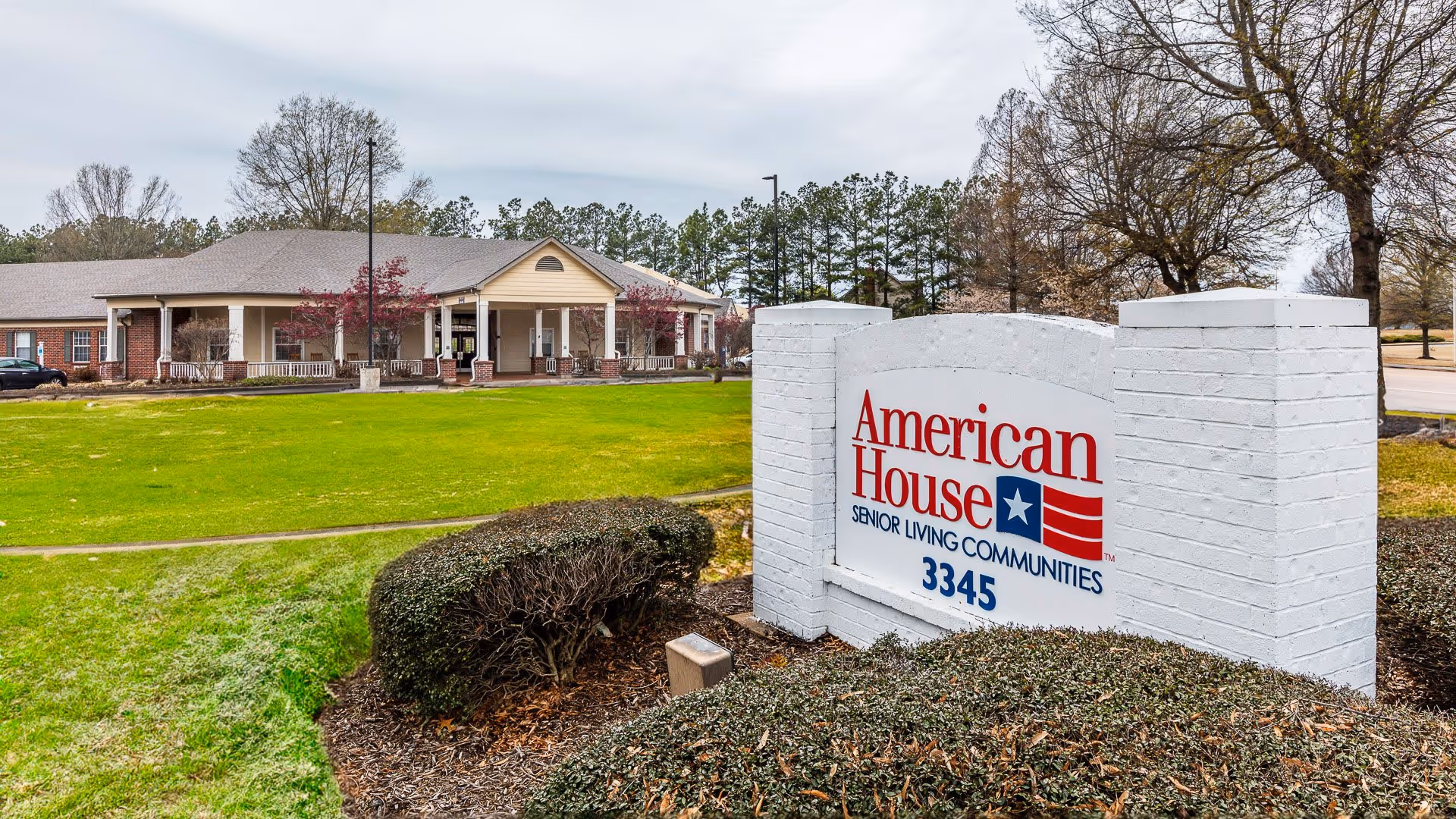 Exterior view of a senior living community building with a large green lawn and trimmed bushes in front. A white brick sign in the foreground reads 'American House Senior Living Communities 3345'. Trees and a cloudy sky are visible in the background.