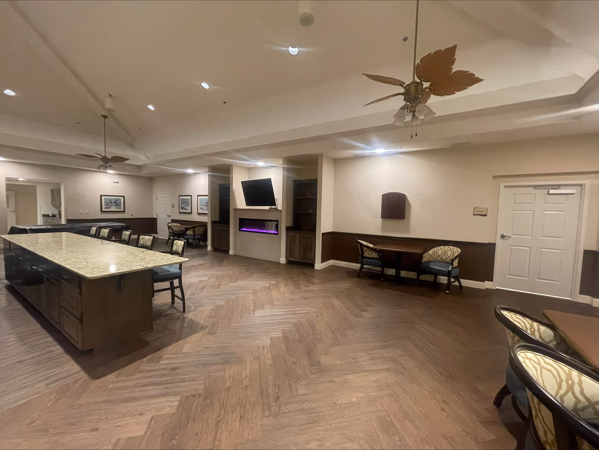Spacious senior living common room with a large island countertop, seating, a wall-mounted TV above an electric fireplace, and ceiling fans.