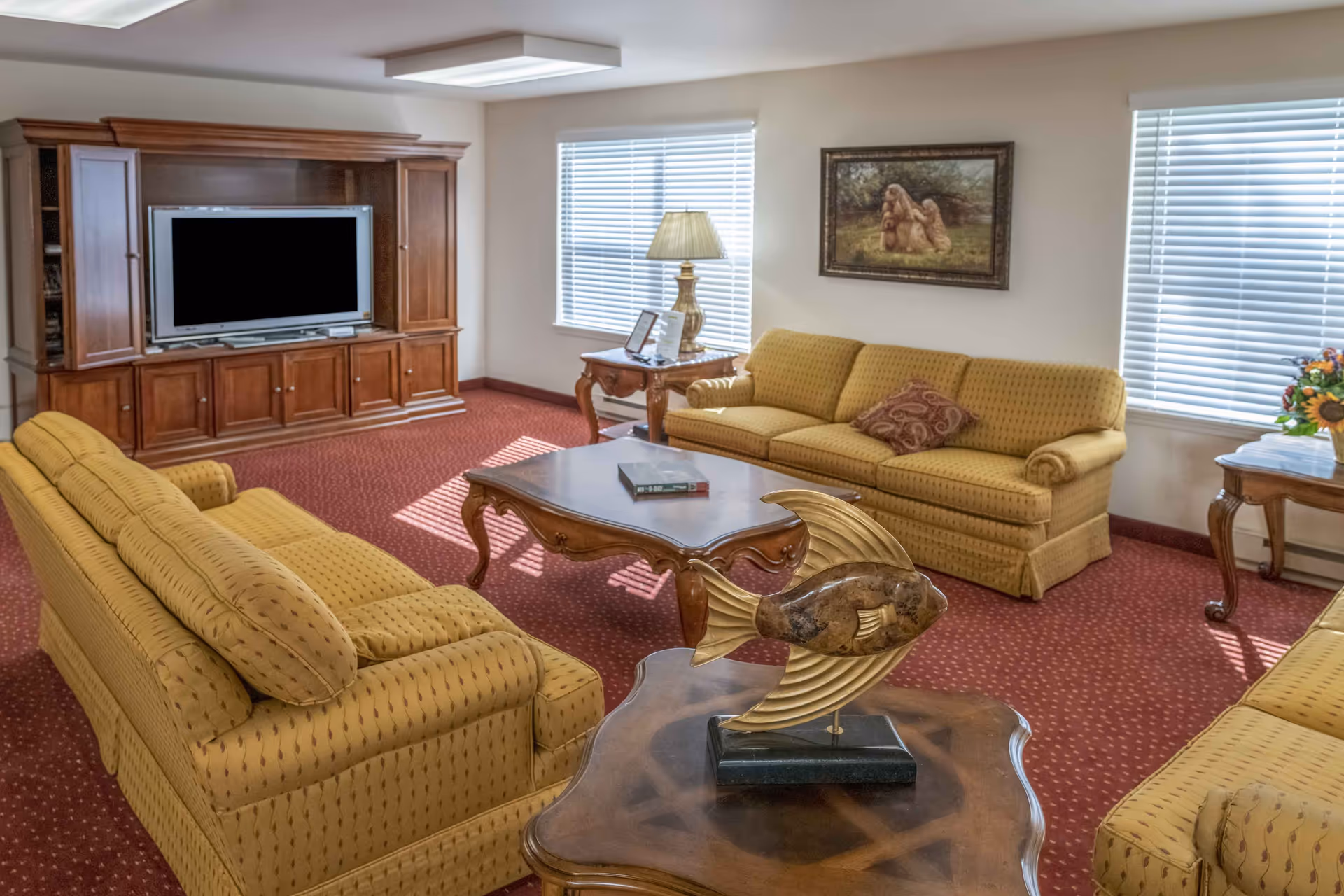 Carpeted community living room with mustard-yellow sofas, a wooden entertainment center and coffee tables with decorative accents.