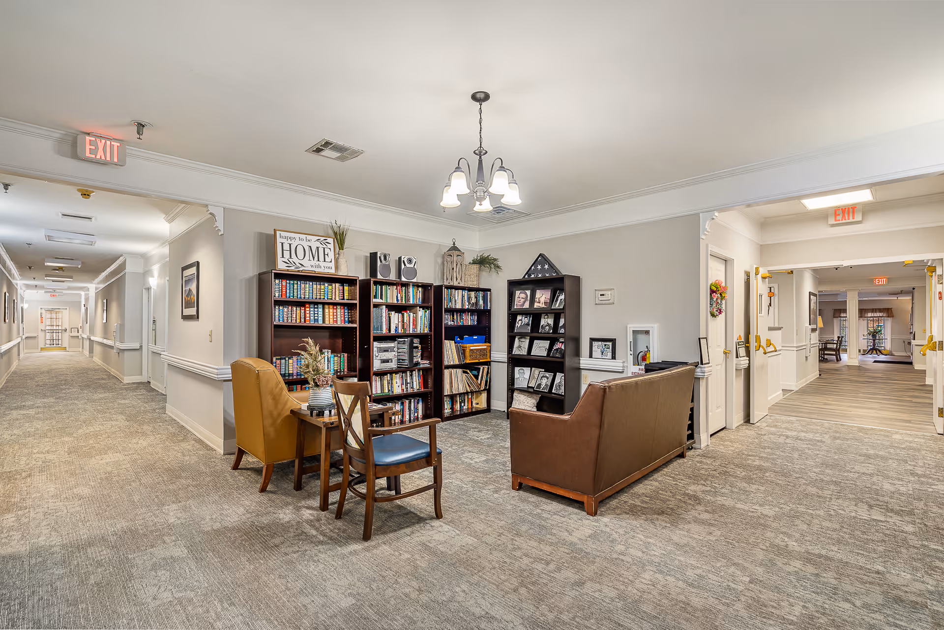 A carpeted senior living common area with bookshelves, chairs, and a sofa flanked by long hallways.