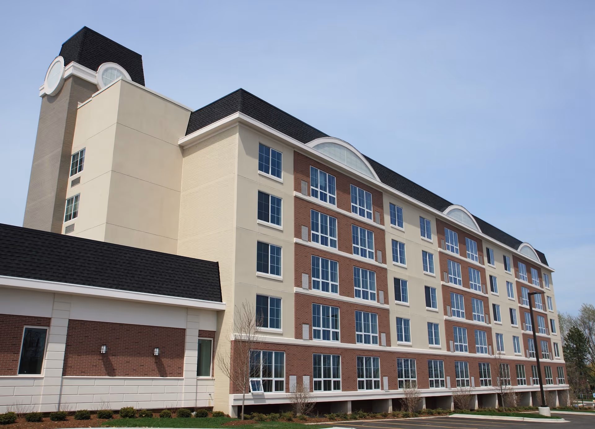 Multi-story brick and stucco senior living building with rows of windows and a dark mansard roof against a clear sky.