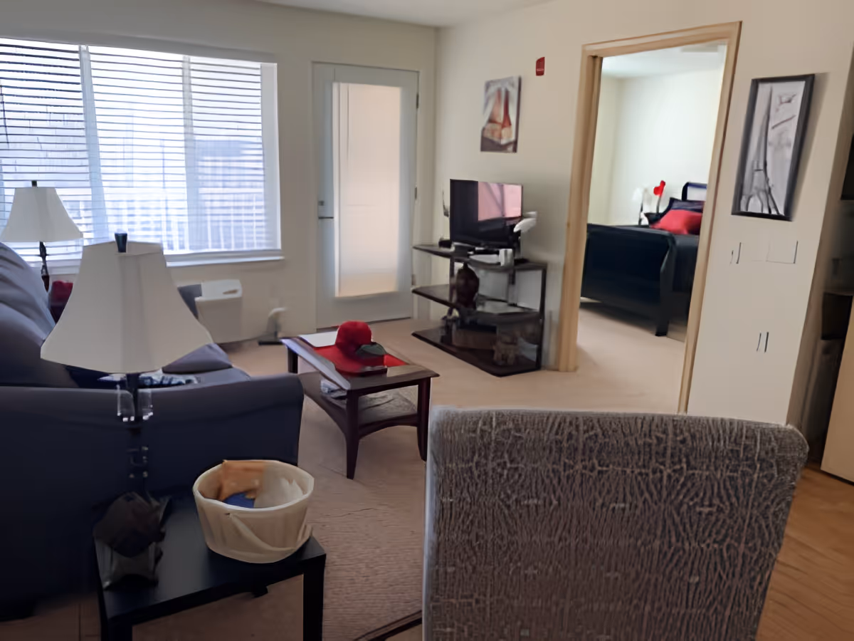 A cozy living room with a gray couch, a side table with a lamp and a basket, a coffee table with a red hat and a book, a TV on a stand, and a doorway leading to a bedroom with a bed and red pillows. Large window with blinds lets in natural light.