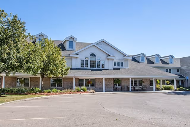 Exterior view of a two-story assisted living facility building with a large parking area in front, several windows, dormer windows on the roof, and a tree on the left side under a clear blue sky.