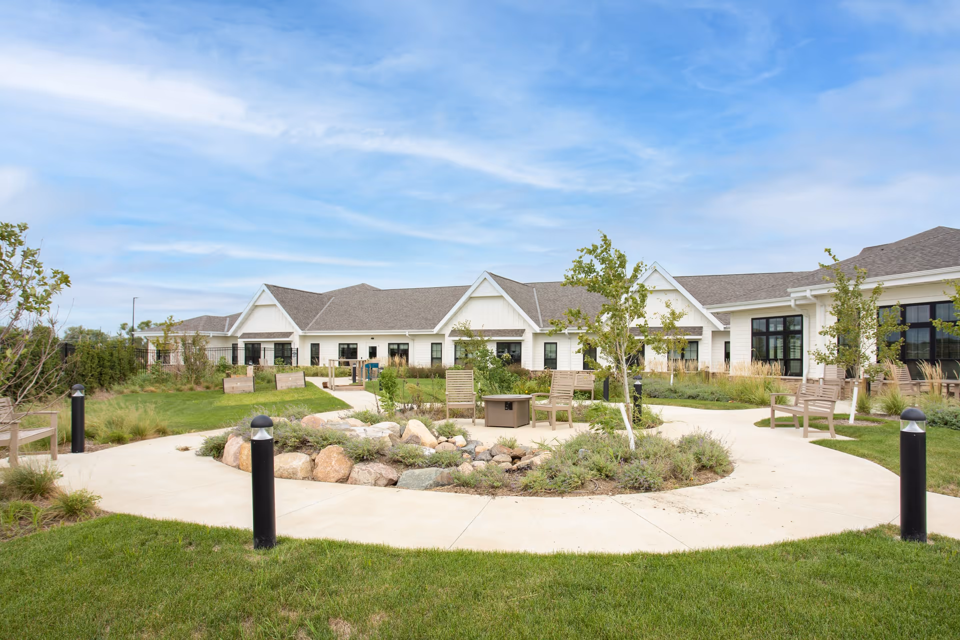 Outdoor garden area at Holland Farms Senior Living featuring a circular concrete pathway surrounding a landscaped rock and plant bed, with several wooden benches and chairs placed around the path. The background shows a single-story building with white siding, multiple windows, and a gray shingled roof under a partly cloudy blue sky.