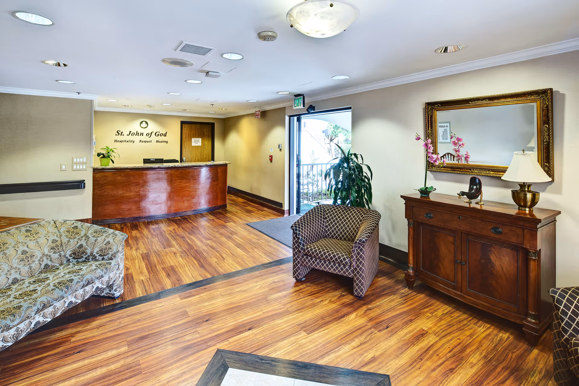 Reception area of St. John of God Retirement & Care Center with a wooden reception desk, a patterned armchair, a floral-patterned sofa, a wooden cabinet with a lamp and decorative items, a large ornate mirror, and a door leading outside.