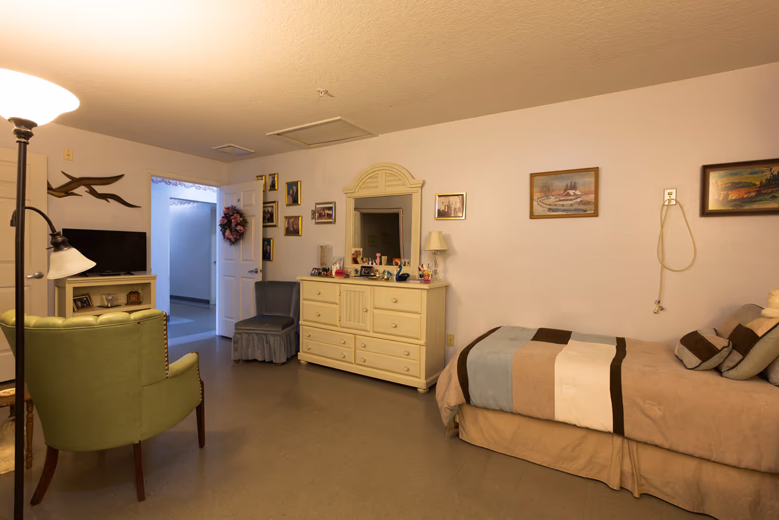 A cozy bedroom in a senior living facility with a single bed covered in a beige, brown, and blue striped blanket. The room has a green armchair, a small TV on a stand, a dresser with a mirror, and several framed pictures on the walls. A door is open, showing a hallway outside the room.