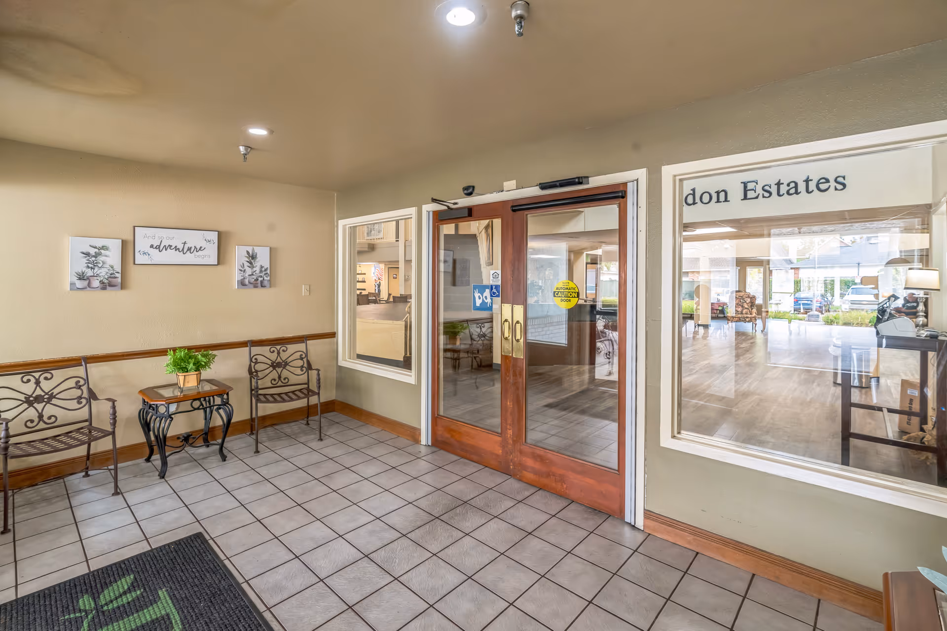 Entrance area of Whealdon Estates by Barclay House featuring double glass doors with wooden frames, a tiled floor, two metal chairs with decorative backs, a small table with a potted plant, and wall art with the phrase 'And so the adventure begins'. Through the glass doors and windows, a spacious interior with seating and large windows is visible.