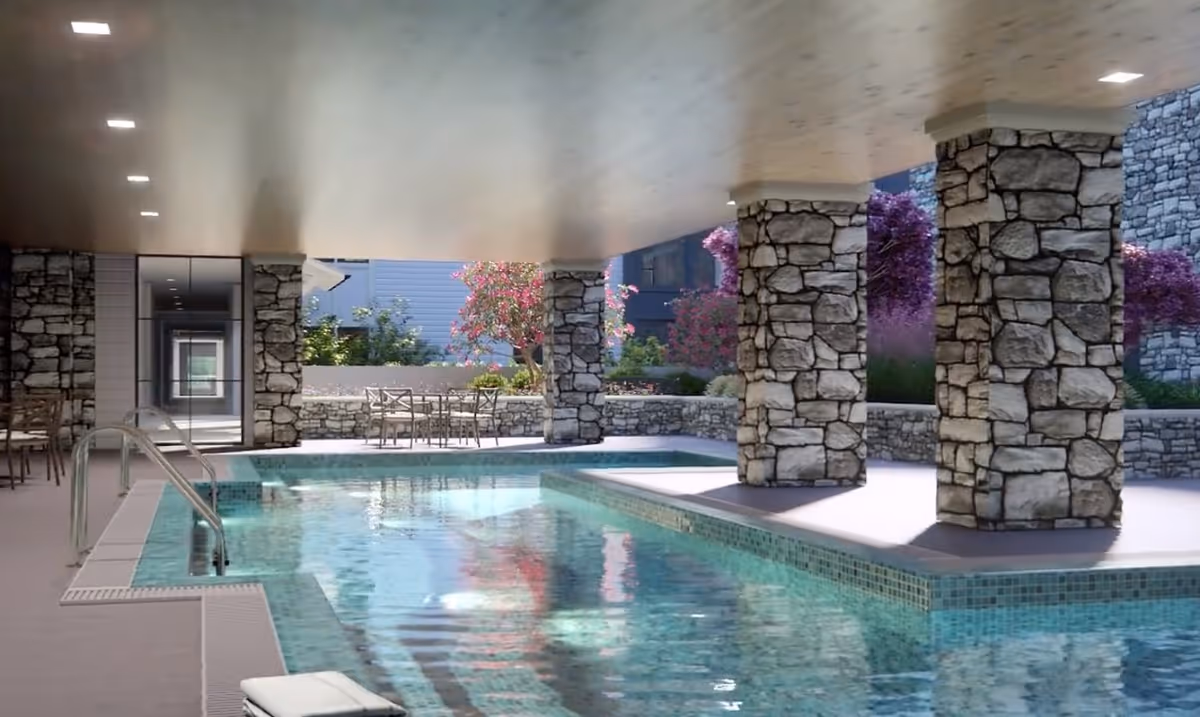 Covered outdoor swimming pool area with stone pillars supporting the ceiling, surrounded by a stone wall and landscaping with flowering bushes and trees. There are metal handrails for pool entry and a few tables and chairs in the background near the garden area.