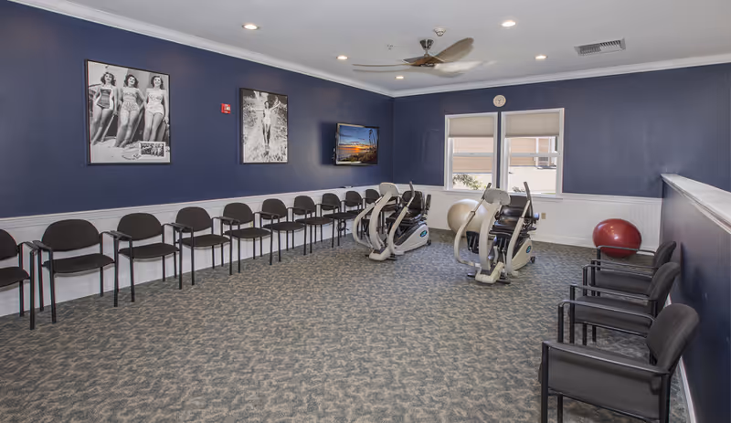 A fitness room with two stationary exercise bikes and two large exercise balls near two windows. The room has blue walls with white wainscoting, a patterned carpet, and a ceiling fan. There are multiple black chairs lined up along two walls, and three black and white vintage-style photos and a flat screen TV mounted on the wall.