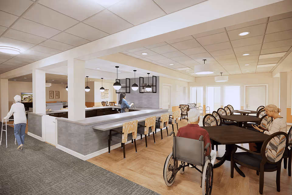 A bright and spacious common area in a senior living facility featuring a kitchen with a long counter and bar stools, several dining tables with chairs, and seniors engaging in various activities. One senior uses a walker, another sits in a wheelchair, and a third is seated at a table looking at a tablet. The room has warm lighting and a welcoming atmosphere.