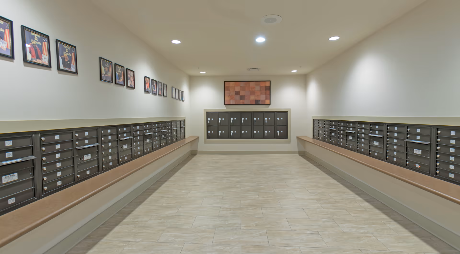 Interior mailroom hallway with rows of resident mailboxes along both walls, bench ledges beneath them and framed artwork on the walls.