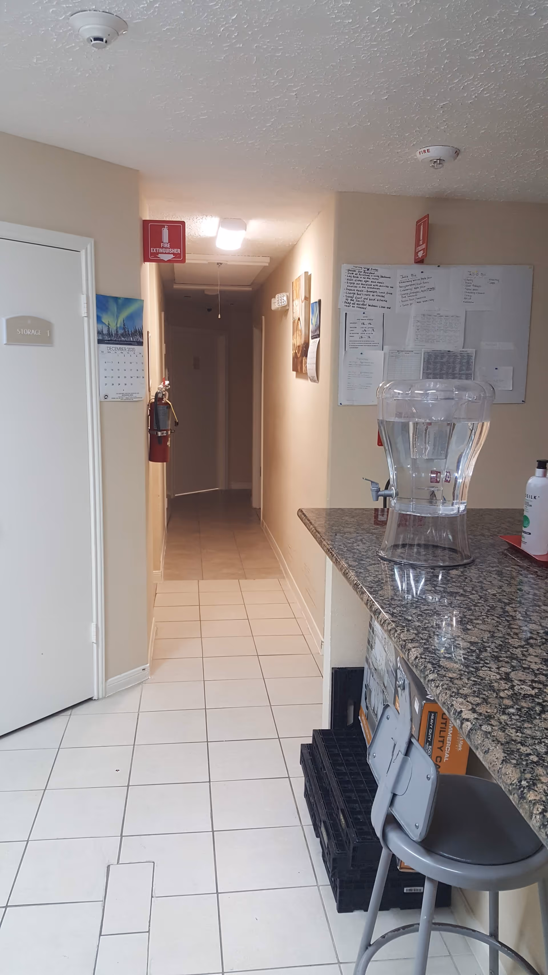 Narrow tiled hallway inside an assisted living facility with a granite countertop and water dispenser in the foreground and a fire extinguisher on the wall.