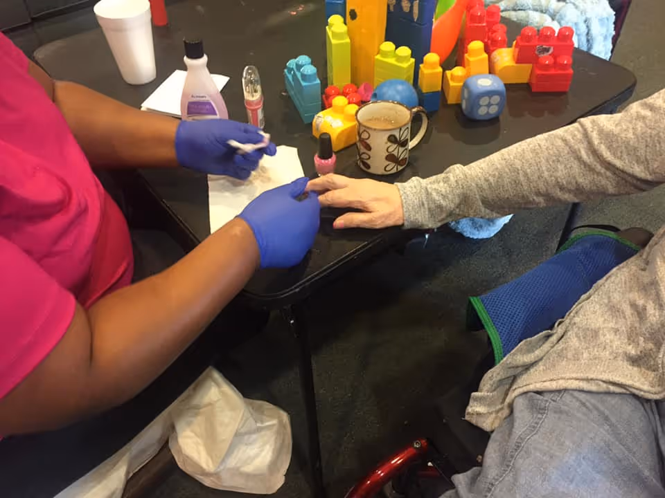 A person wearing blue gloves is applying pink nail polish to the fingernails of an elderly person seated at a table. The table has colorful building blocks, a coffee mug, and nail care items on it. The elderly person is wearing a gray long-sleeve shirt and sitting in a wheelchair.