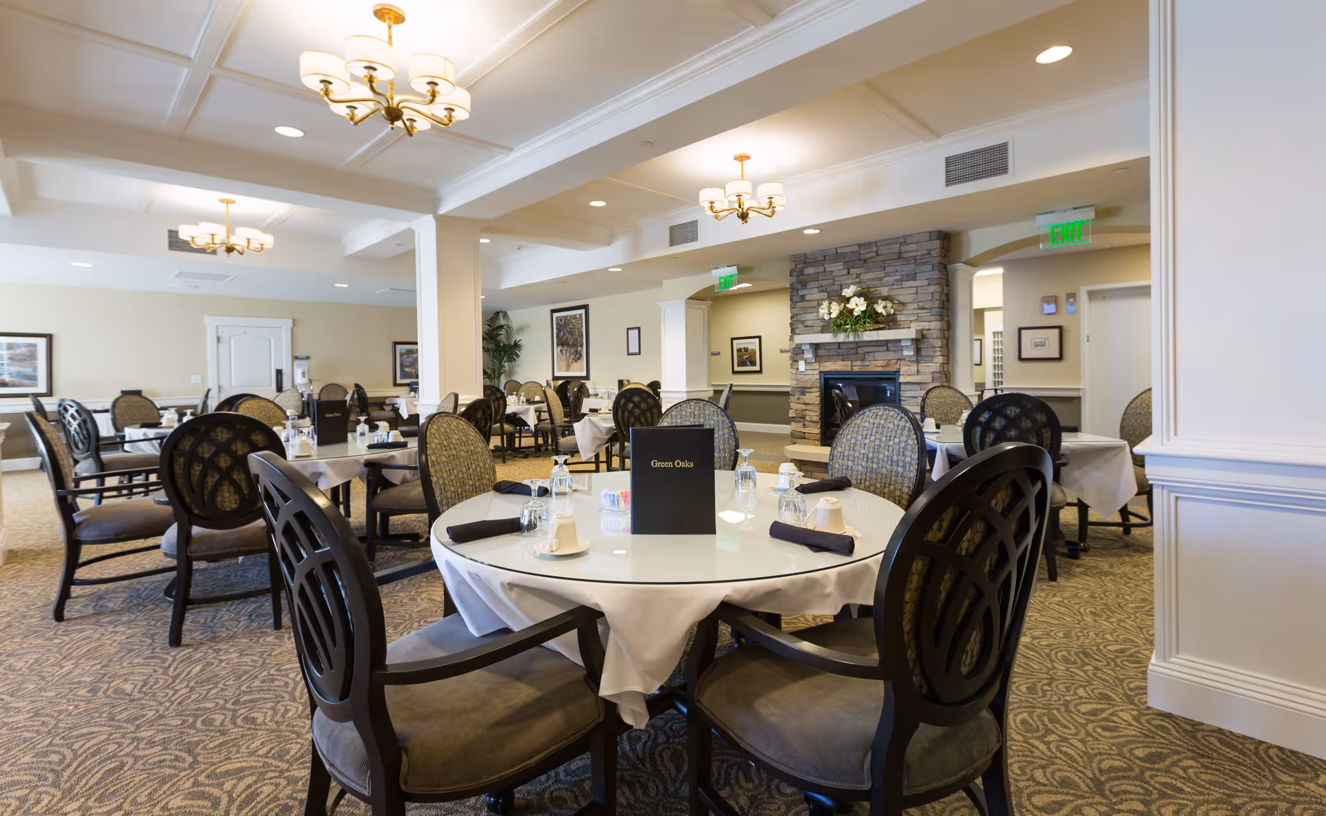 A spacious dining room in a senior living facility with round tables covered in white tablecloths, set with glasses, napkins, and cups. The room features elegant chairs, a stone fireplace with floral decoration, framed artwork on the walls, and warm lighting from ceiling chandeliers.