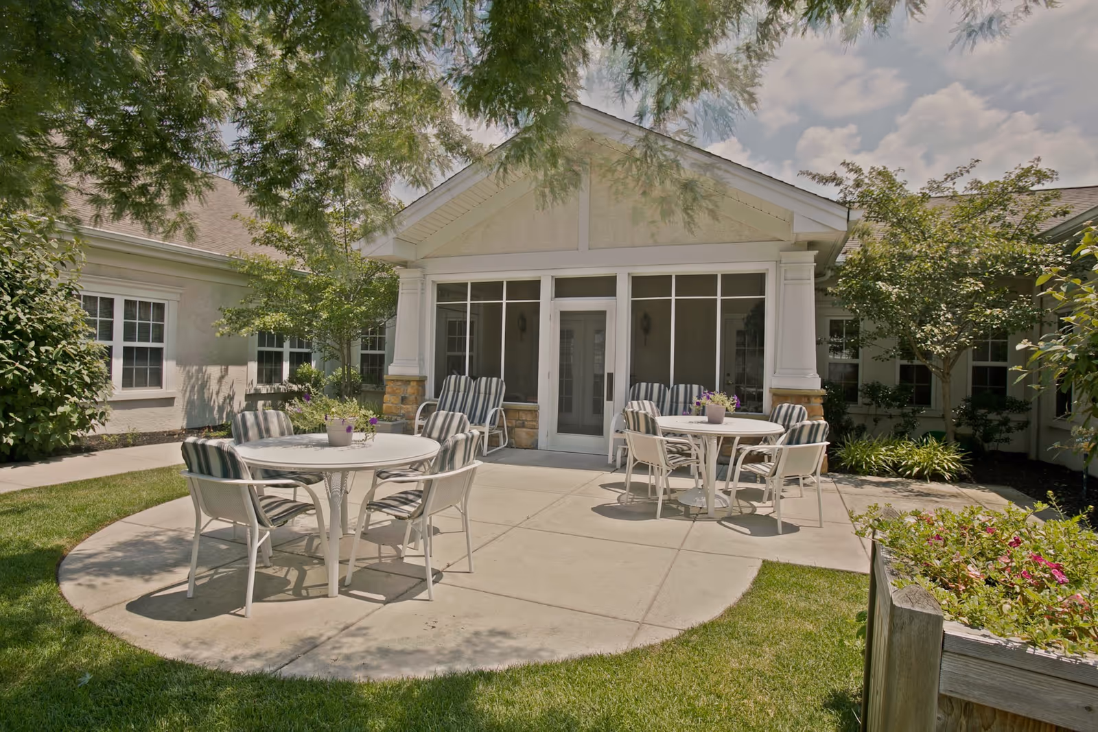 Outdoor patio area at Brandywine Reflections by Monarch with two round tables surrounded by striped cushioned chairs, potted plants on the tables, and a building entrance with large windows and a glass door in the background. Trees and greenery surround the patio under a partly cloudy sky.