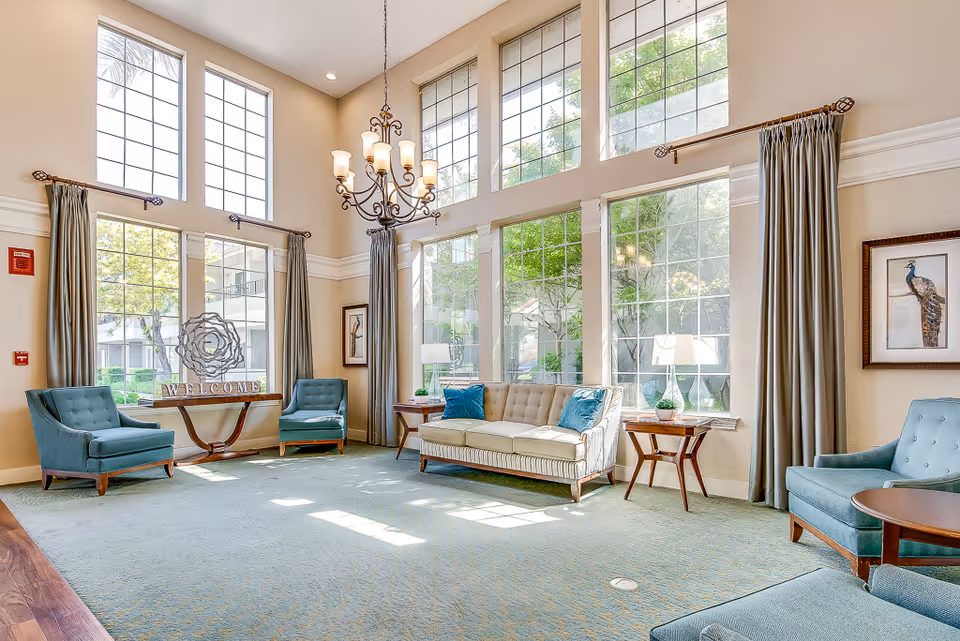 A bright and spacious senior living facility common area with large floor-to-ceiling windows letting in natural light. The room features a beige sofa with blue cushions, several teal armchairs, wooden side tables with lamps, a chandelier, and a decorative 'WELCOME' sign on a console table. The walls are light-colored with framed artwork and long curtains.