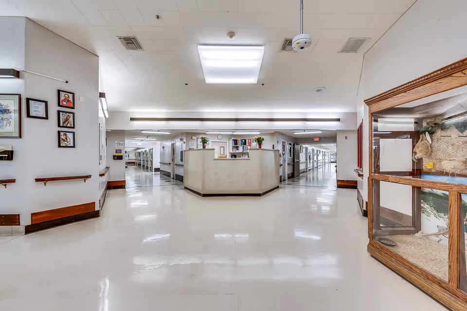 A wide, clean hallway in a nursing center with a central nurse's station. The hallway has white walls and floors with wooden handrails along the sides. There are framed pictures on the left wall and a glass display case with a taxidermy animal on the right. The ceiling has bright fluorescent lighting.