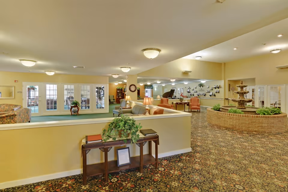 A spacious senior living facility common area with patterned carpet flooring, beige walls, and multiple seating arrangements including sofas and armchairs. There is a brick circular fountain with greenery in the center right, a wooden table with plants and books in the foreground, and glass-paneled doors leading to another room in the background. The area is well-lit with ceiling lights.