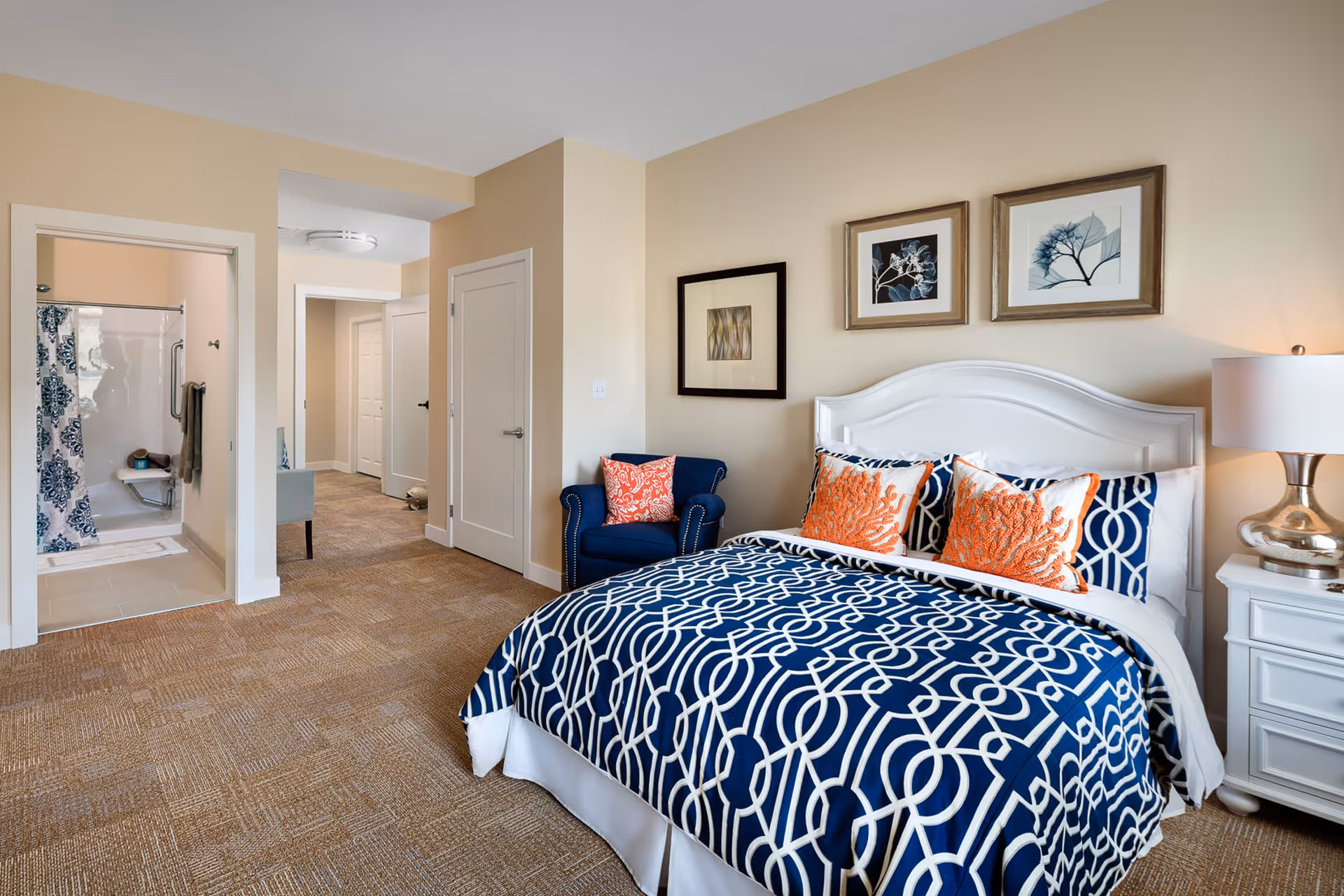 A bedroom with a white bed frame and a blue and white patterned bedspread with orange decorative pillows. There is a blue armchair with an orange pillow next to the bed. On the wall above the bed are three framed artworks. To the right of the bed is a white nightstand with a silver lamp. The room has beige walls and carpeted floor. An open doorway leads to a bathroom with a shower that has a blue and white patterned curtain.