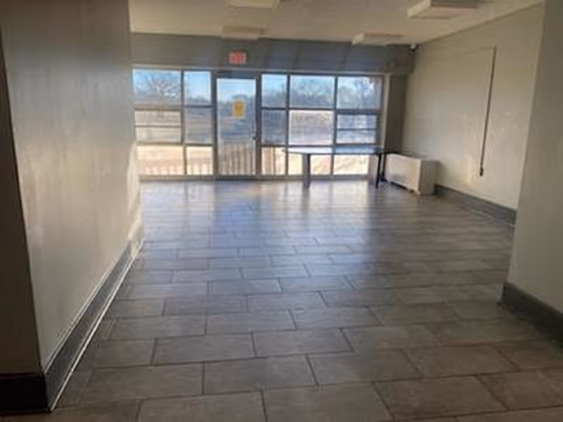 Empty room with tiled floor and large windows on one side letting in natural light. There is a door in the center of the windows and a small radiator or heater unit against the right wall.