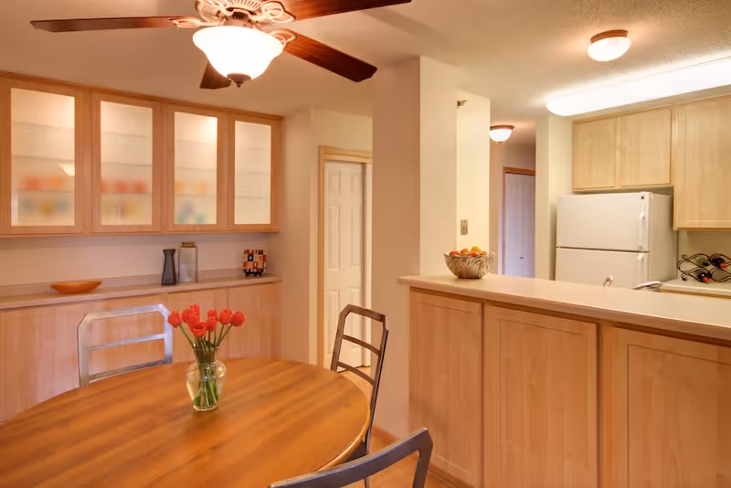 A cozy dining area with a round wooden table and metal chairs. A vase with red tulips is placed on the table. Behind the table, there is a wooden cabinet with glass doors displaying blurred items inside. To the right, a kitchen area is visible with light wood cabinets, a white refrigerator, and a small wine rack. The ceiling has a fan with a light fixture.