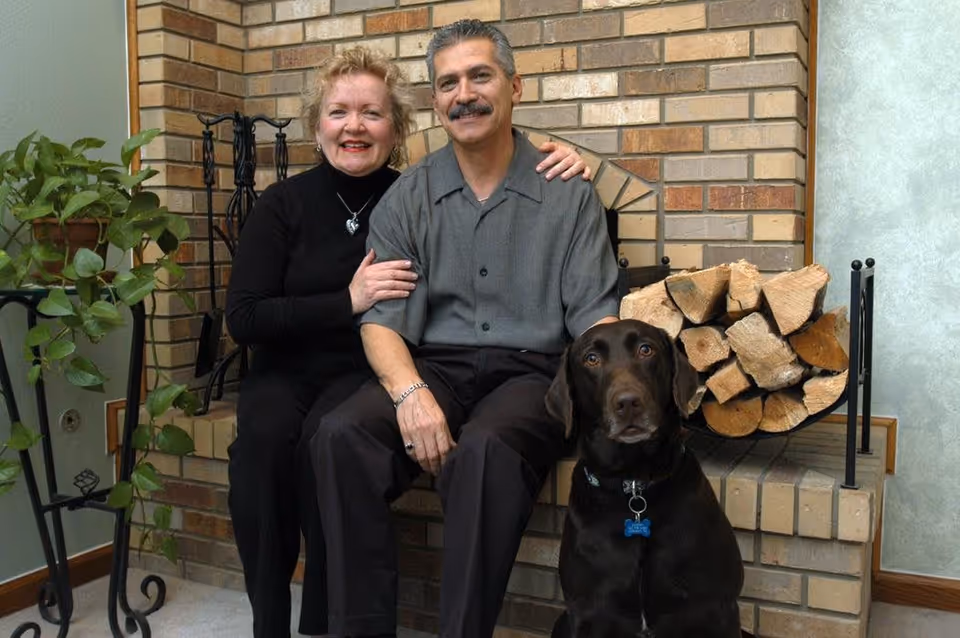A smiling middle-aged couple sitting together in front of a brick fireplace with a stack of firewood. A large brown dog with a blue bone-shaped tag sits in front of them. There is a green potted plant on a black metal stand to the left.