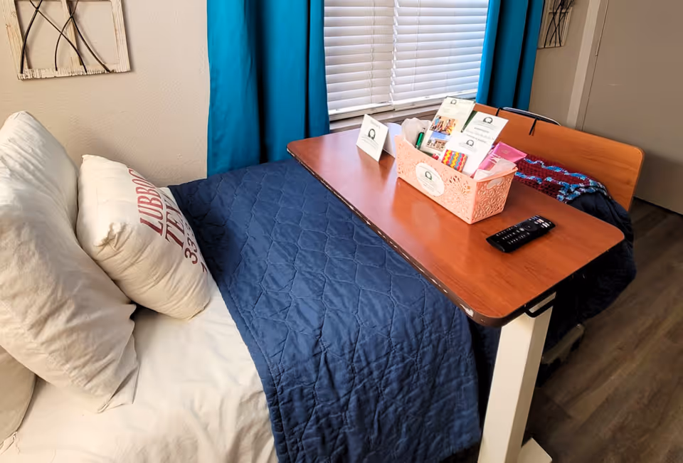 A single bed in a senior facility room with a navy quilt, pillows, an overbed table holding a basket of brochures and a TV remote, and teal curtains at a window.