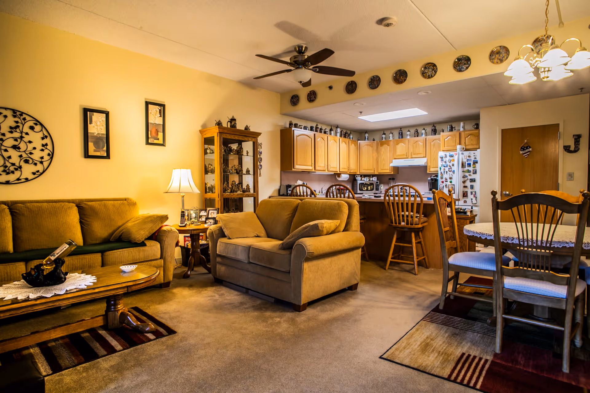 A cozy living room and kitchen area in a senior living facility. The living room has two beige sofas, a wooden coffee table with a decorative centerpiece, a side table with a lamp and framed photos, and wall decorations. The kitchen features wooden cabinets, a refrigerator covered with magnets, a microwave, and a counter with wooden bar stools. A dining table with chairs is visible on the right side under a chandelier light fixture.