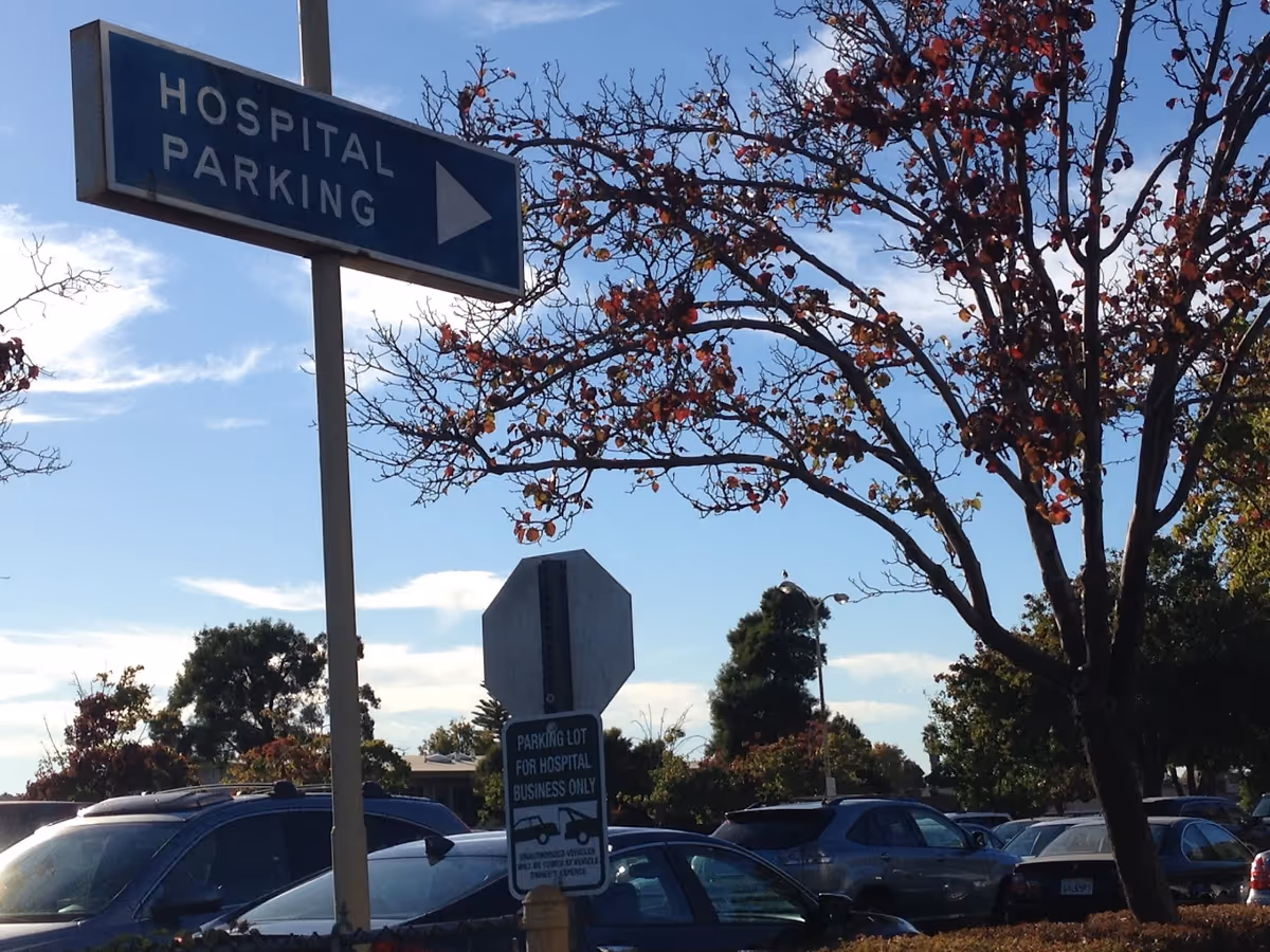 Parking lot with a 'Hospital Parking' sign, several parked cars, and trees under a blue sky.