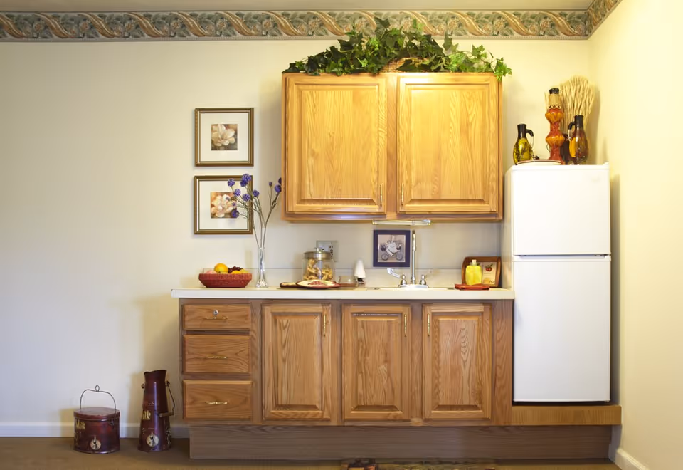 A small kitchen area with wooden cabinets, a white refrigerator, a countertop with a sink, decorative items including a vase with purple flowers, framed pictures on the wall, and ornamental bottles on top of the refrigerator. There are also two vintage milk containers on the floor to the left.