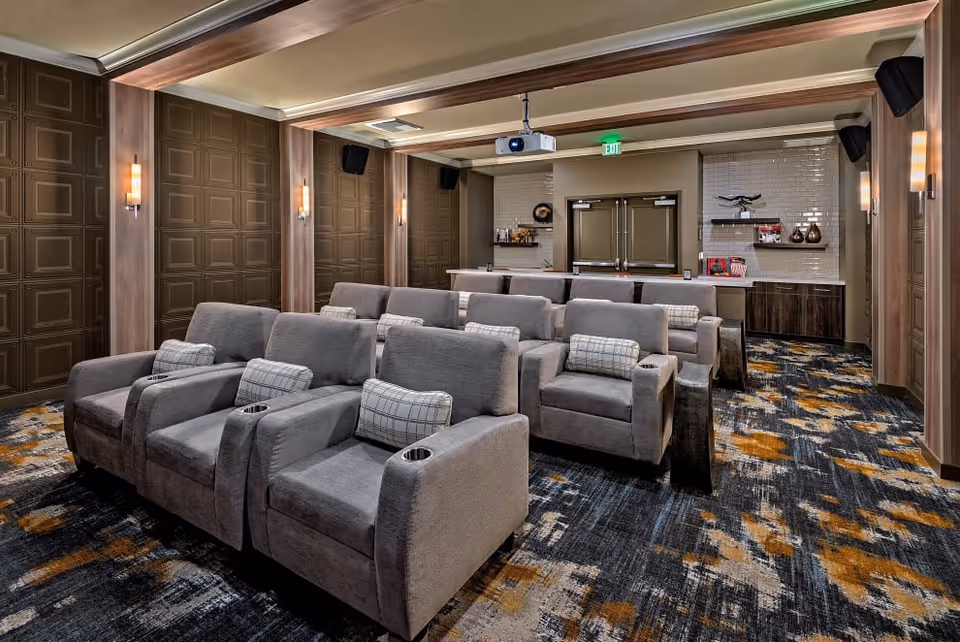 A cozy theater room with two rows of gray reclining seats, each equipped with cup holders and plaid pillows. The room features patterned carpet with shades of gray, blue, and orange, paneled walls with sconces, a ceiling-mounted projector, and a counter area at the back with shelves and double doors.
