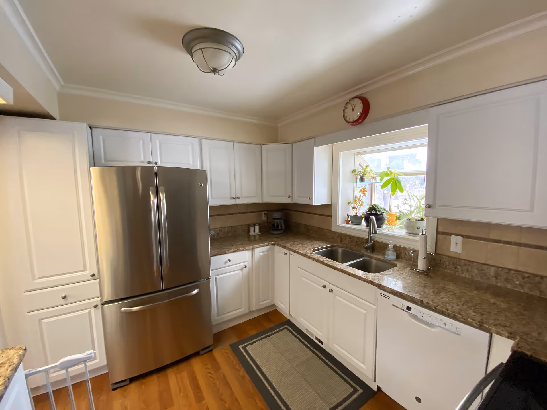 A clean kitchen with white cabinets, a stainless steel refrigerator, granite countertops, a double sink under a window with several potted plants, a dishwasher, and a wall clock above the window. The floor is wooden with a rectangular rug in front of the sink.